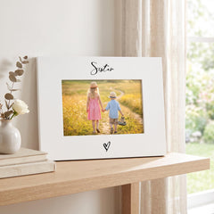 A white picture frame with a photograph of two children, a girl and a boy, standing on a path in a field. The word "Sister" is written above the photograph.