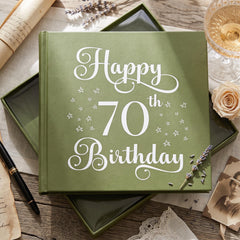 A green book titled "Happy 70th Birthday" sits on a wooden surface, surrounded by various objects such as a glass, a rose, and some papers.