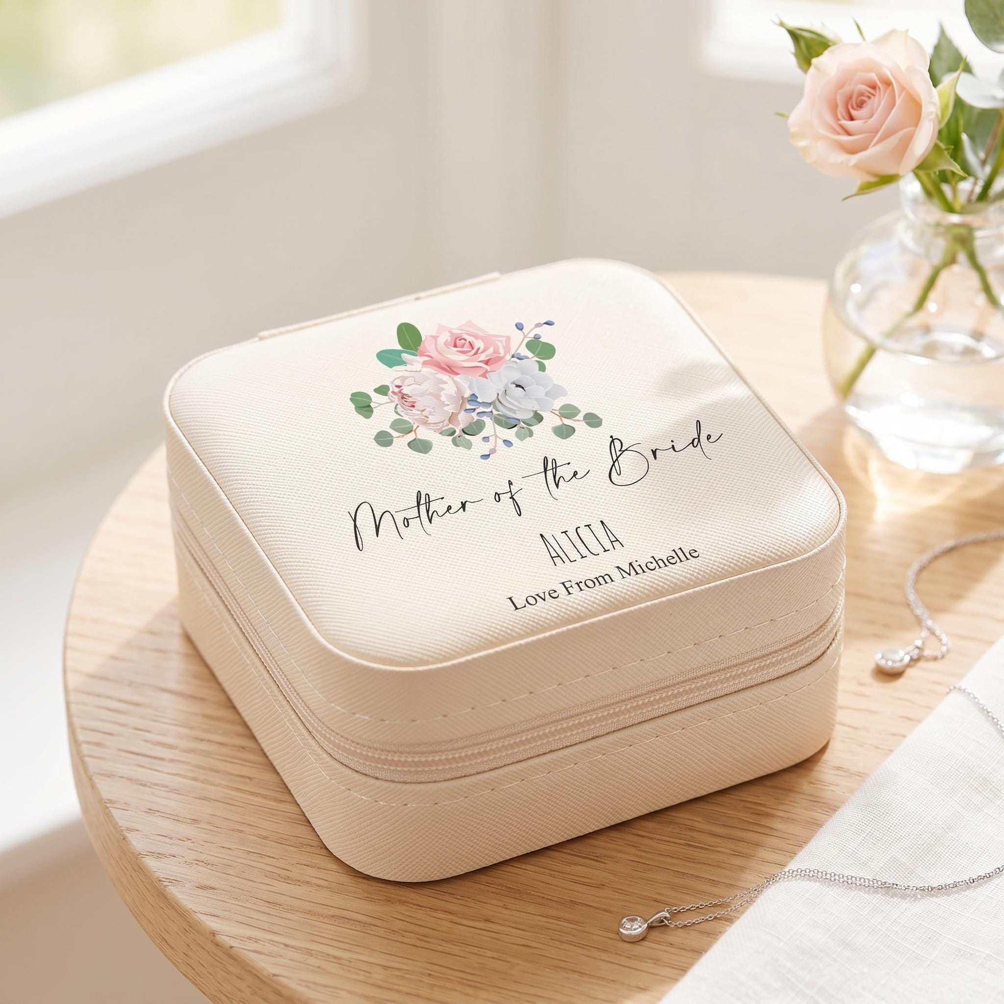 A white jewelry case with a floral design and the text "Mother of the Bride" printed on it, placed on a wooden surface next to a vase with roses.