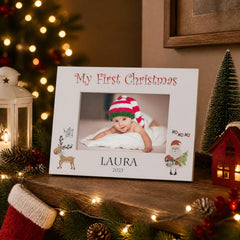 A white picture frame with a photo of a baby wearing a red and white striped hat, surrounded by Christmas decorations and lights.