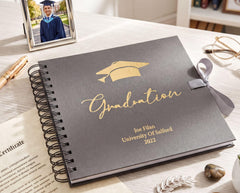 a graduation album with a graduation cap on top, surrounded by graduation-related items such as a graduation cap, a graduation gown, a graduation certificate, a pen, a pair of glasses, and a succulent plant.