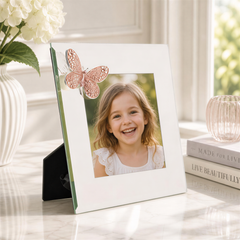 A young girl with a bright smile is the focus of a white picture frame with a butterfly decoration, placed on a marble surface next to a vase of white flowers and a stack of books.