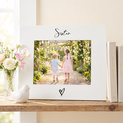 A white picture frame with a photo of two children, one boy and one girl, standing in a garden surrounded by flowers. The frame has the word "Sister" written on it. The frame is placed on a wooden shelf next to a vase of flowers and a small white bird figurine.