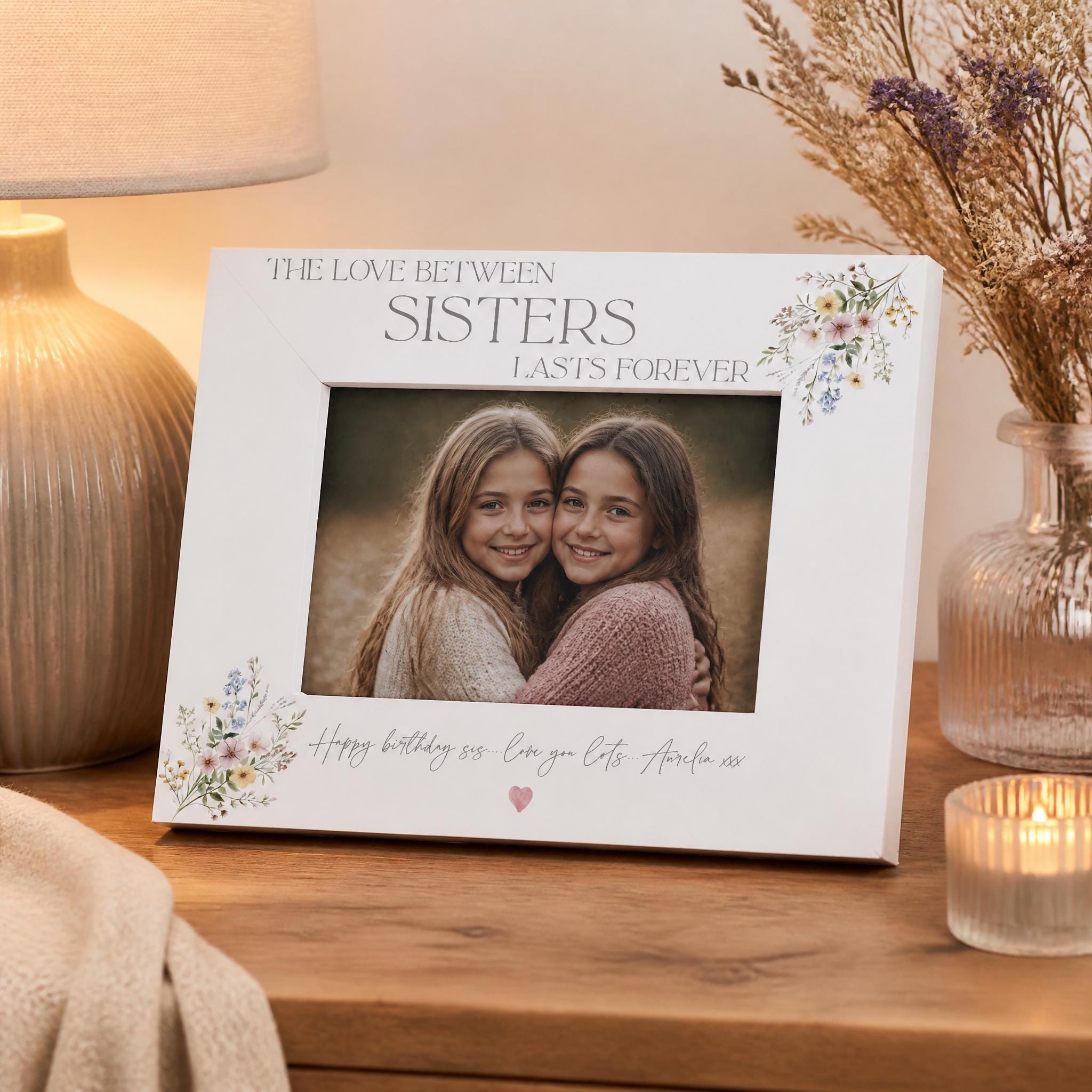 A white picture frame with a photo of two smiling young girls inside, placed on a wooden surface. The frame has a floral design and the text "The love between sisters lasts forever" written on it.