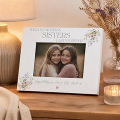 A white picture frame with a photo of two smiling young girls inside, placed on a wooden surface. The frame has a floral design and the text "The love between sisters lasts forever" written on it.