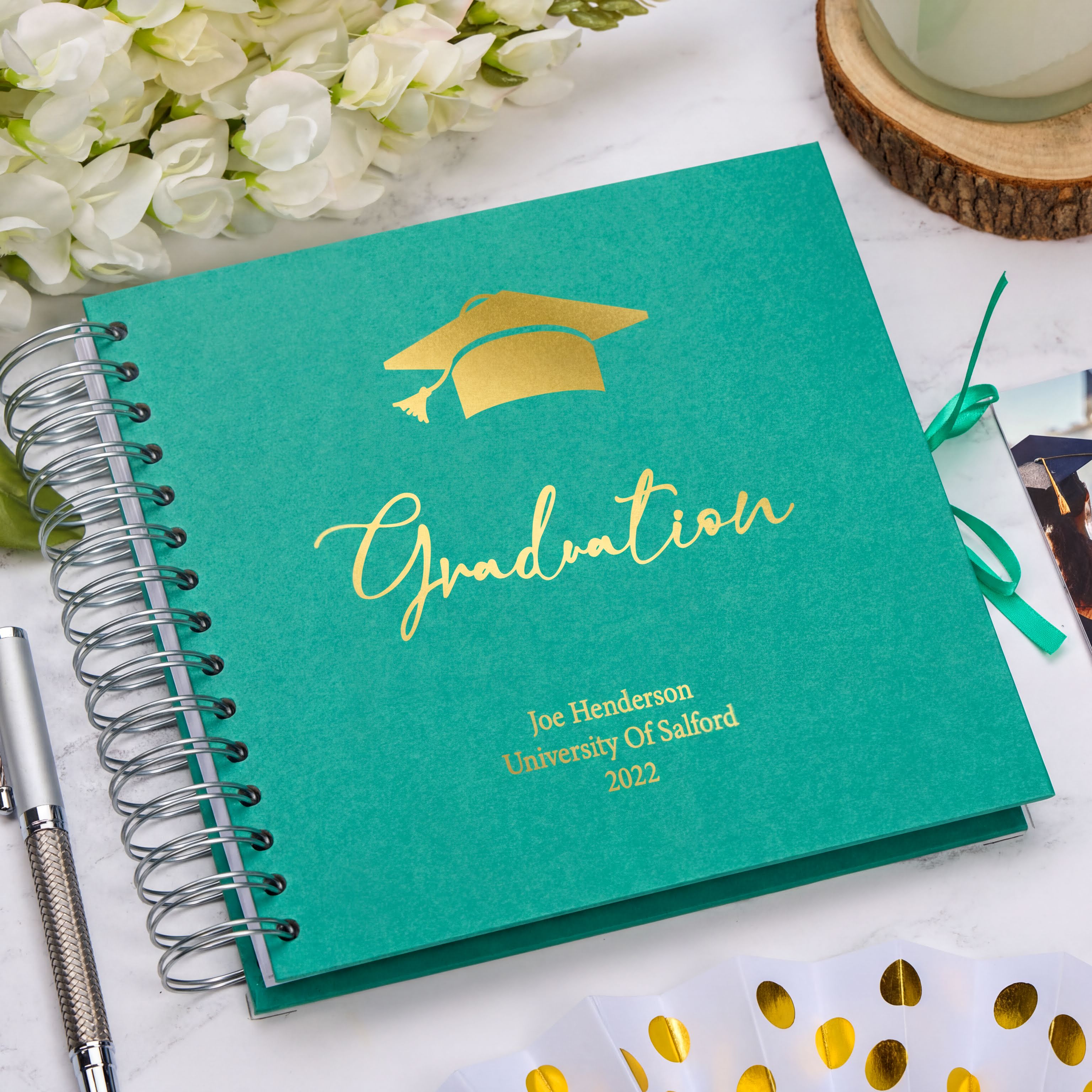 A graduation cap-themed spiral-bound notebook with the text "Graduation" and "Joe Henderson University Of Salford 2020" printed on it, placed on a white surface alongside a pen, flowers, and a wooden coaster.