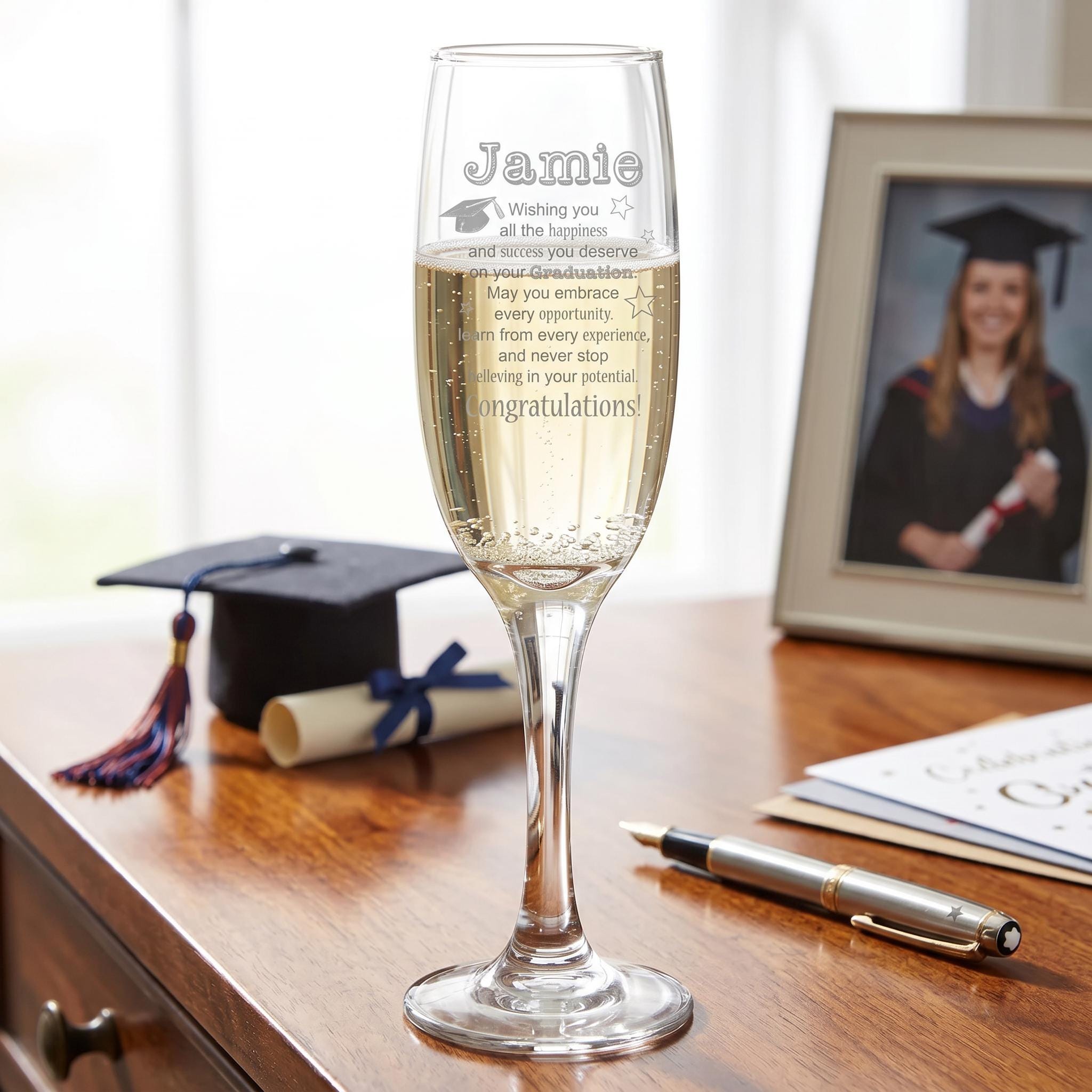 A personalized graduation glass with a heartfelt message, placed on a wooden desk next to a graduation cap, a pen, and some papers.