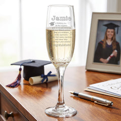 A personalized graduation glass with a heartfelt message, placed on a wooden desk next to a graduation cap, a pen, and some papers.