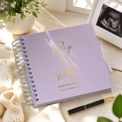 A purple baby shower book titled "Bump to Baby" sits on a wooden surface, accompanied by a pen and a framed image of an ultrasound.