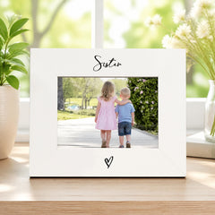 A white picture frame with a photo of two children, a girl and a boy, standing together in a garden. The word "Sister" is written above the photo.