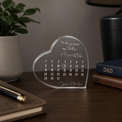 A transparent heart-shaped calendar with a personalized message sits on a wooden desk, surrounded by a potted plant, a notebook, and a book.