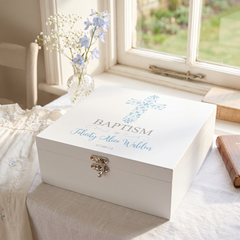 A white wooden box with a blue cross design and the text "Baptism" is placed on a table next to a vase of white flowers and a book.