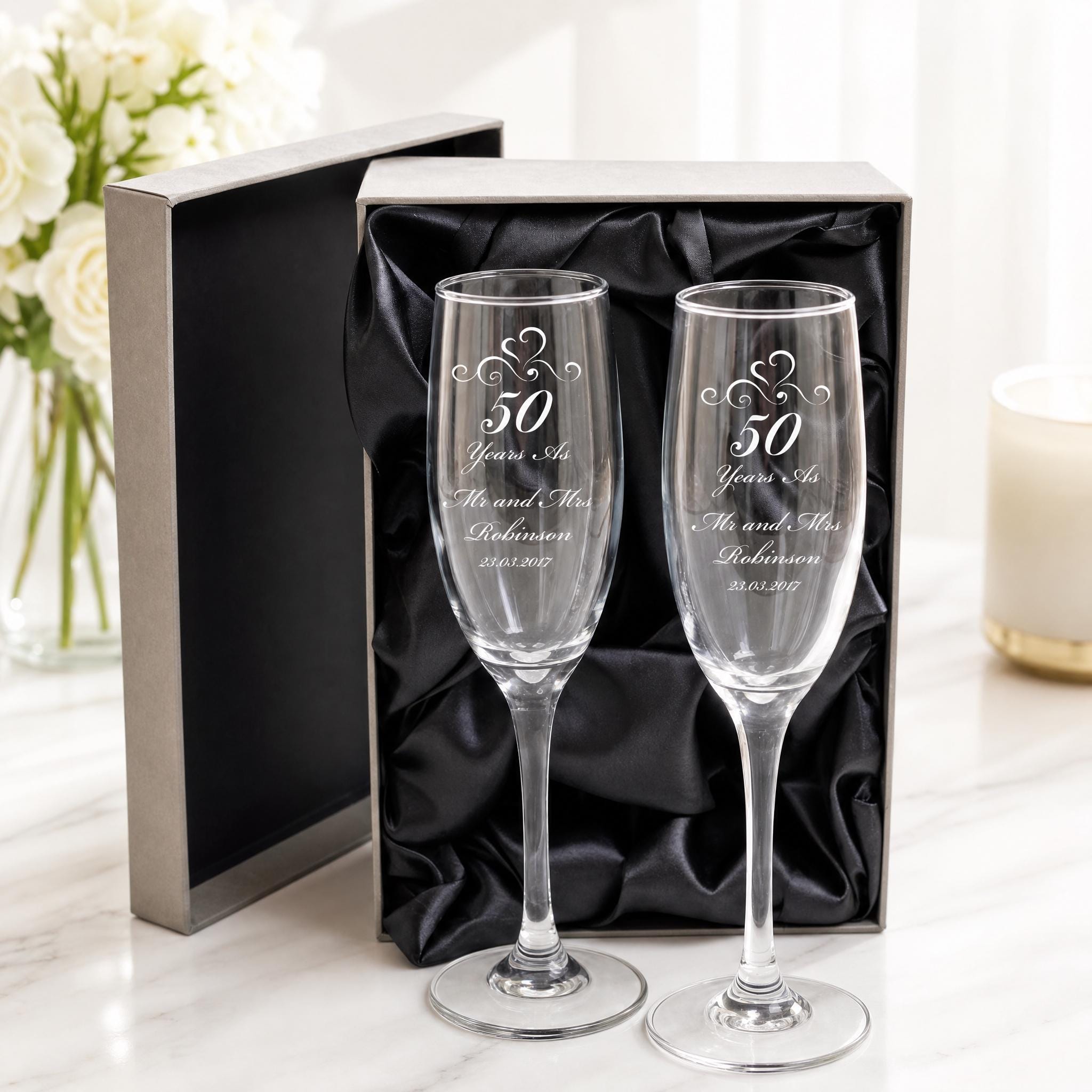 a pair of elegant champagne glasses, each engraved with a message celebrating a 50th anniversary. The glasses are placed in a black box, which is placed on a marble surface. In the background, there is a vase filled with white flowers, adding a touch of elegance to the scene.