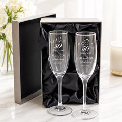 a pair of elegant champagne glasses, each engraved with a message celebrating a 50th anniversary. The glasses are placed in a black box, which is placed on a marble surface. In the background, there is a vase filled with white flowers, adding a touch of elegance to the scene.