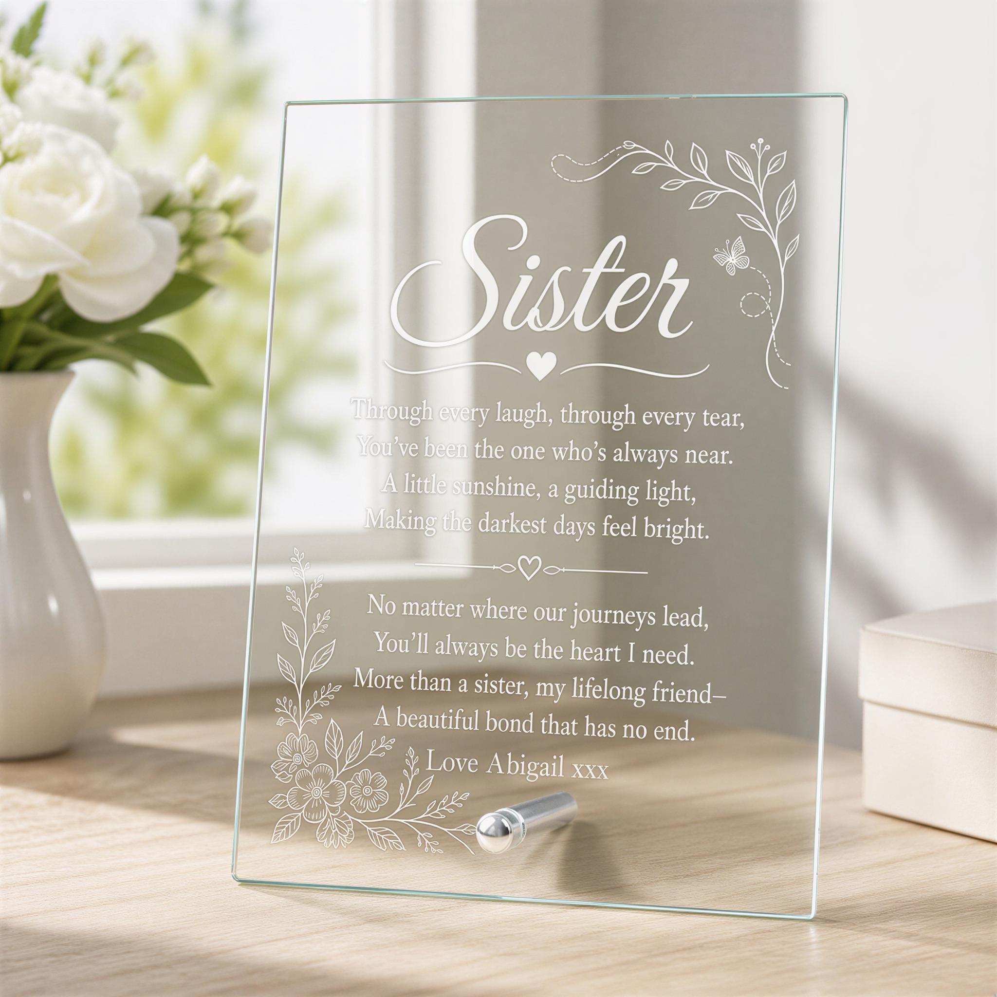 A glass frame with a heartfelt message about a sister, placed on a wooden surface next to a vase of white flowers.