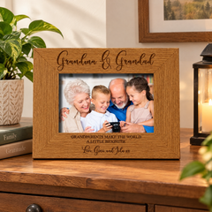 A wooden picture frame with a family photo of a group of people, including a young boy, an elderly couple, and their children, sitting together and smiling. The frame has a message engraved on it that reads "Grandma & Grandpa make the world a little brighter".