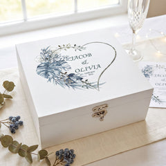 A white wooden box with a heart-shaped design and floral elements, placed on a table with a glass of wine and some flowers.