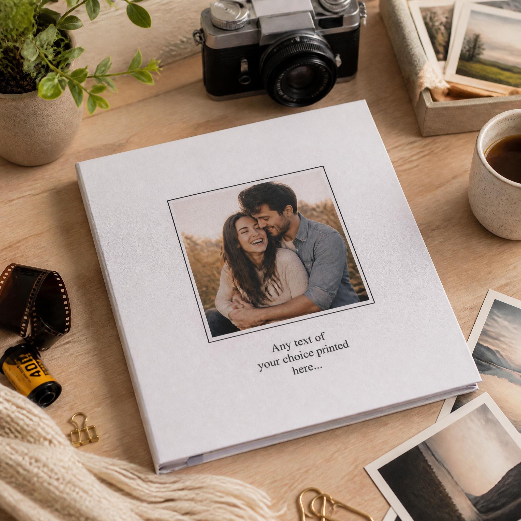 A white photo album with a printed photo of a couple embracing is placed on a wooden surface, surrounded by various items such as a camera, a mug, and some plants.
