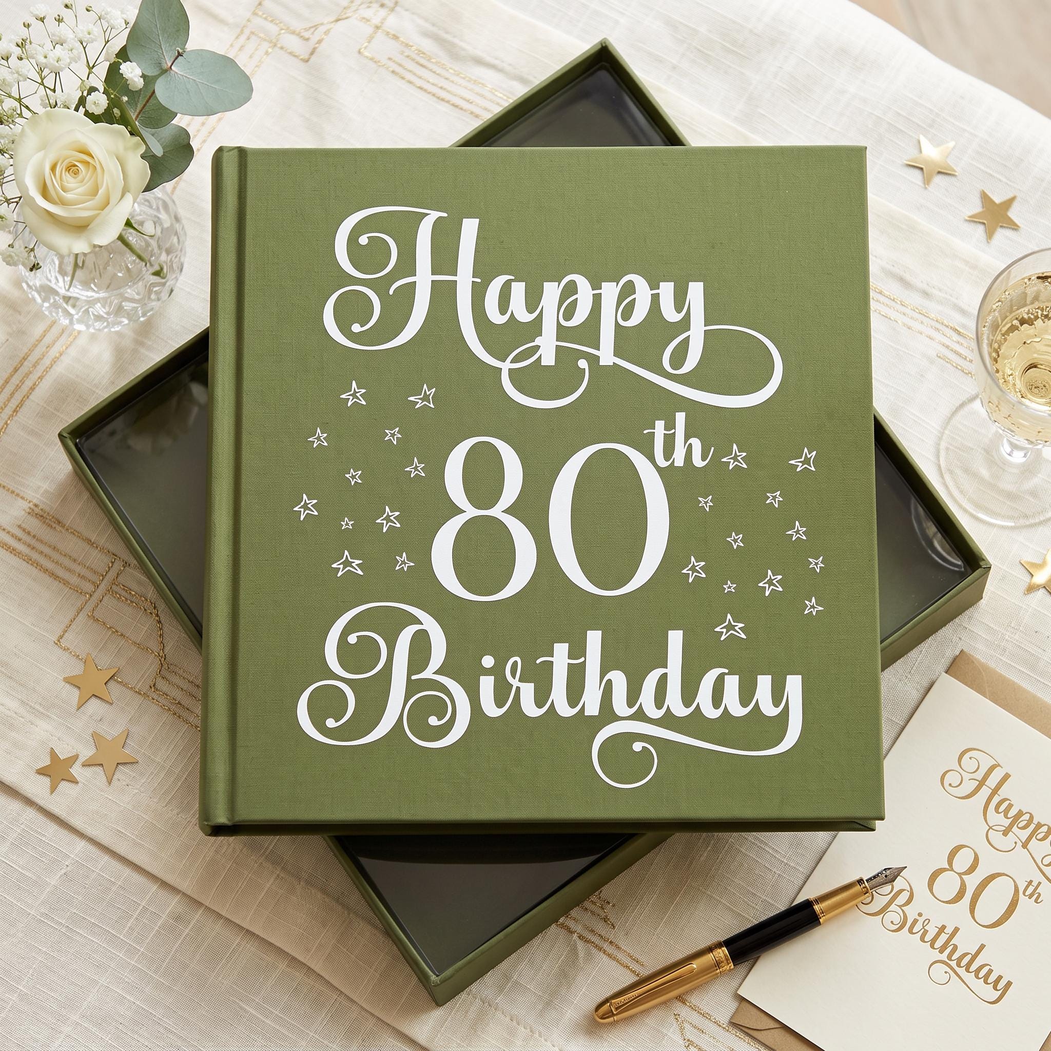 A green book titled "Happy 80th Birthday" sits on a table, surrounded by a vase of white flowers, a glass of champagne, and gold stars.