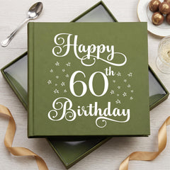 A green photo album with the text "Happy 60th Birthday" printed on it, placed on a wooden surface alongside a glass of champagne and a gold ribbon.