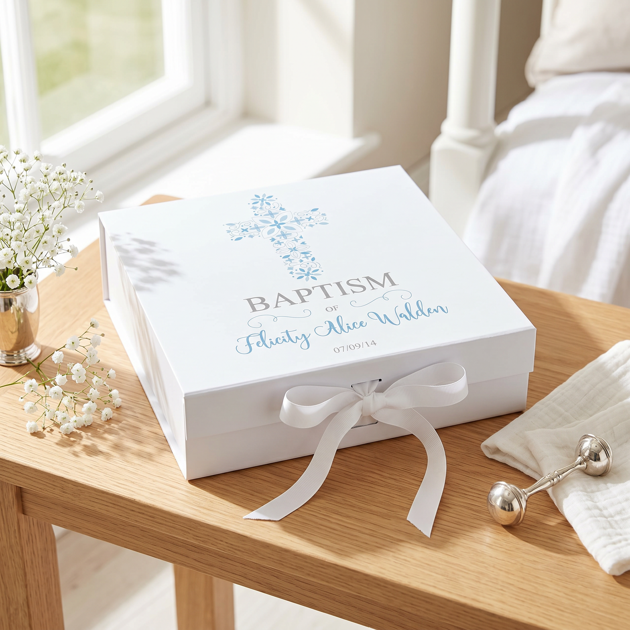 A white box with a blue cross and the text "Baptism" is placed on a wooden table, accompanied by a vase of white flowers and a white towel.