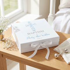 A white box with a blue cross and the text "Baptism" is placed on a wooden table, accompanied by a vase of white flowers and a white towel.
