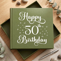 A green book titled "Happy 50th Birthday" with white text and stars on the cover, placed on a wooden surface with some rocks and greenery in the background.