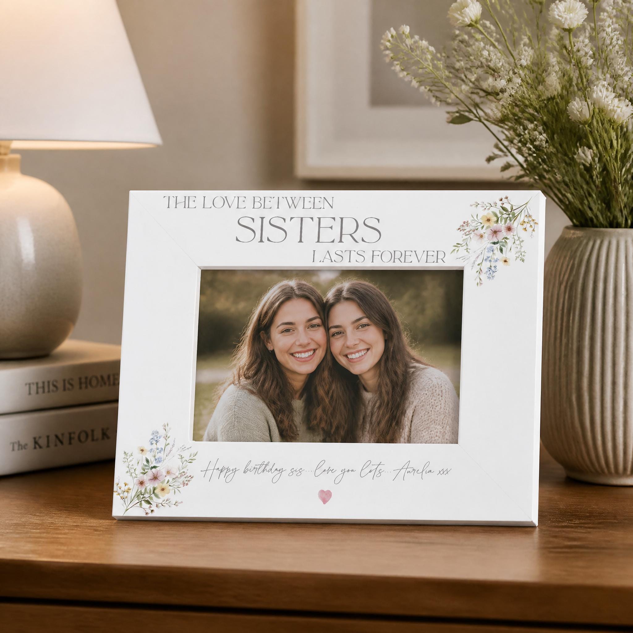 a white picture frame with a photo of two smiling women inside, placed on a wooden surface next to a vase of flowers and a lamp.
