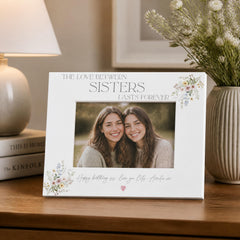 a white picture frame with a photo of two smiling women inside, placed on a wooden surface next to a vase of flowers and a lamp.