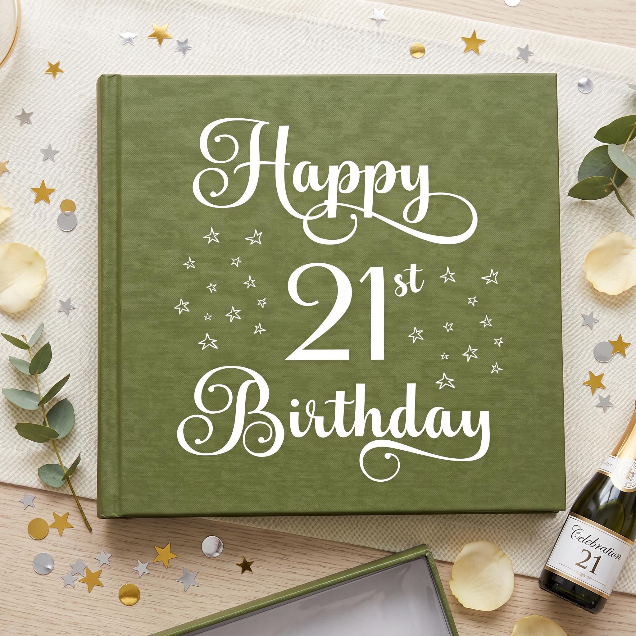 a green book with the text "Happy 21st Birthday" written on it, surrounded by confetti, a bottle of champagne, and some leaves.