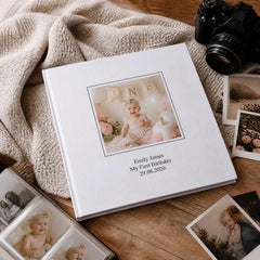 a white photo album with a photograph of a baby girl, placed on a wooden surface alongside a camera and other photographs.