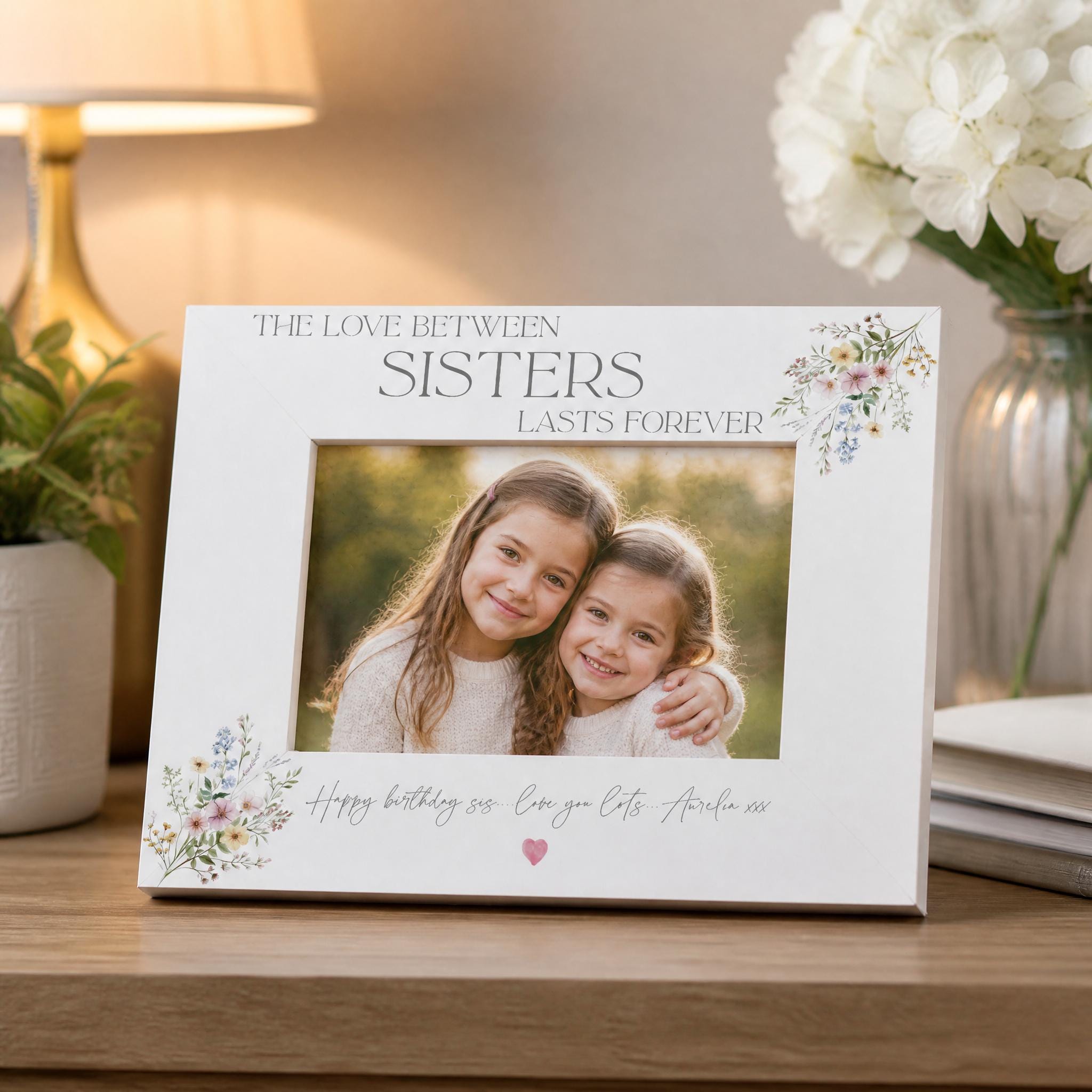 A white picture frame with a photo of two young girls inside, placed on a wooden surface. The frame has a floral design and the text "The love between sisters lasts forever" written on it.