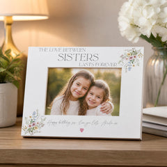A white picture frame with a photo of two young girls inside, placed on a wooden surface. The frame has a floral design and the text "The love between sisters lasts forever" written on it.