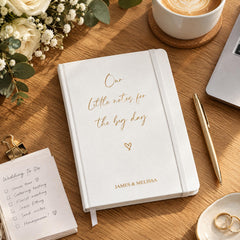 a white notebook with gold text, a pen, a coffee cup, a bouquet of white flowers, and a ring on a wooden table.