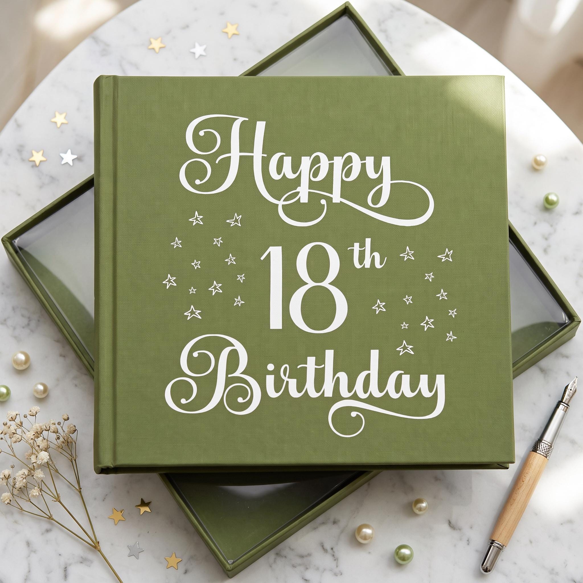 A green book titled "Happy 18th Birthday" with white stars on the cover, placed on a marble surface with scattered pearls and a pen.