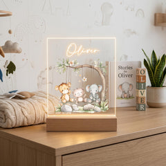 A lighted display case containing a picture of three cartoon animals, including a lion, a bear, and an elephant, along with the name "Oliver" written on it. The display case is placed on a wooden surface, and there are books and a plant nearby.
