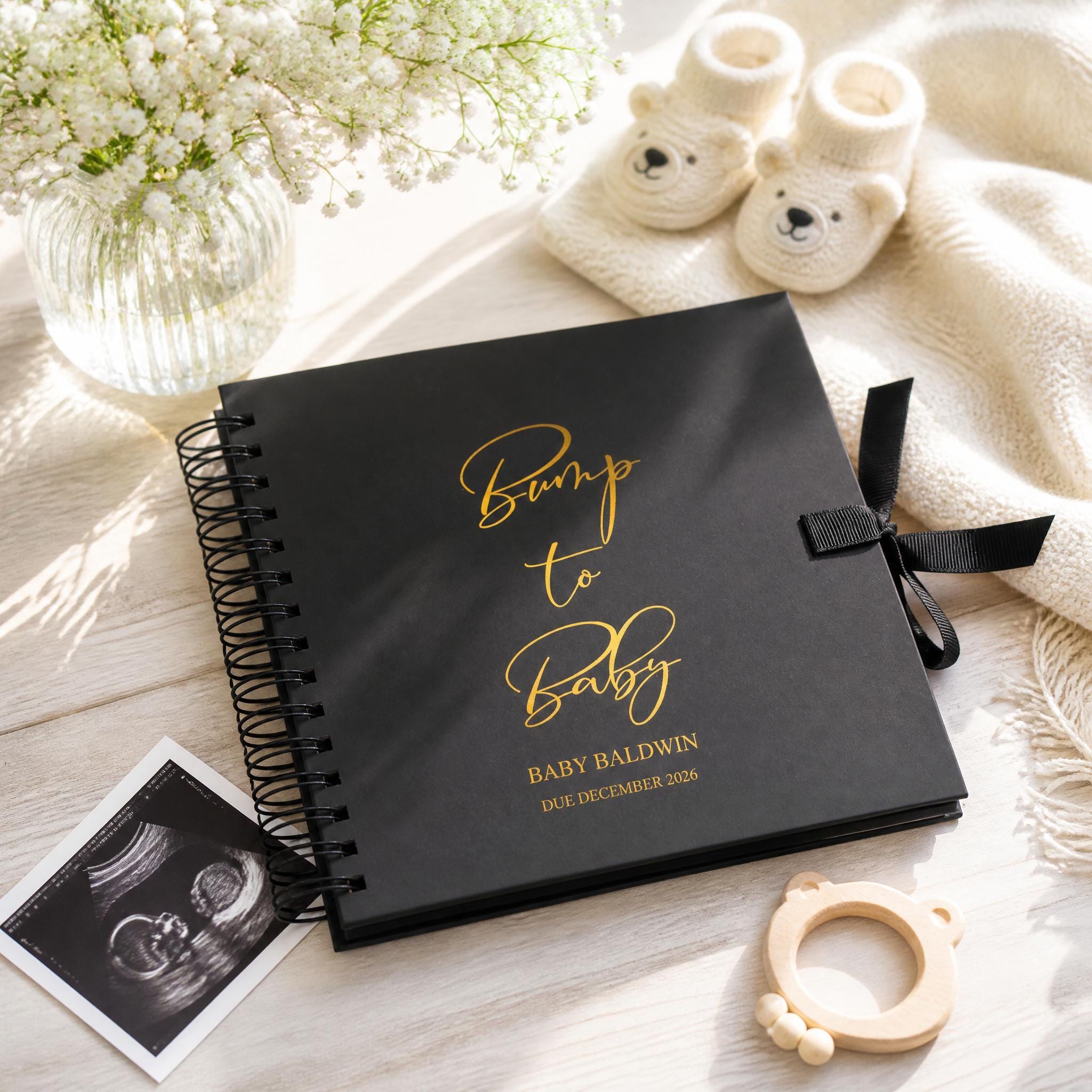 a black photo album with gold text, a vase of white flowers, a pair of white baby booties, and a black and white photograph of a baby's head.
