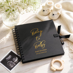 a black photo album with gold text, a vase of white flowers, a pair of white baby booties, and a black and white photograph of a baby's head.