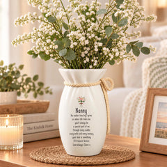 A white vase with a floral arrangement sits on a woven mat, accompanied by a framed picture and a candle on a wooden table.