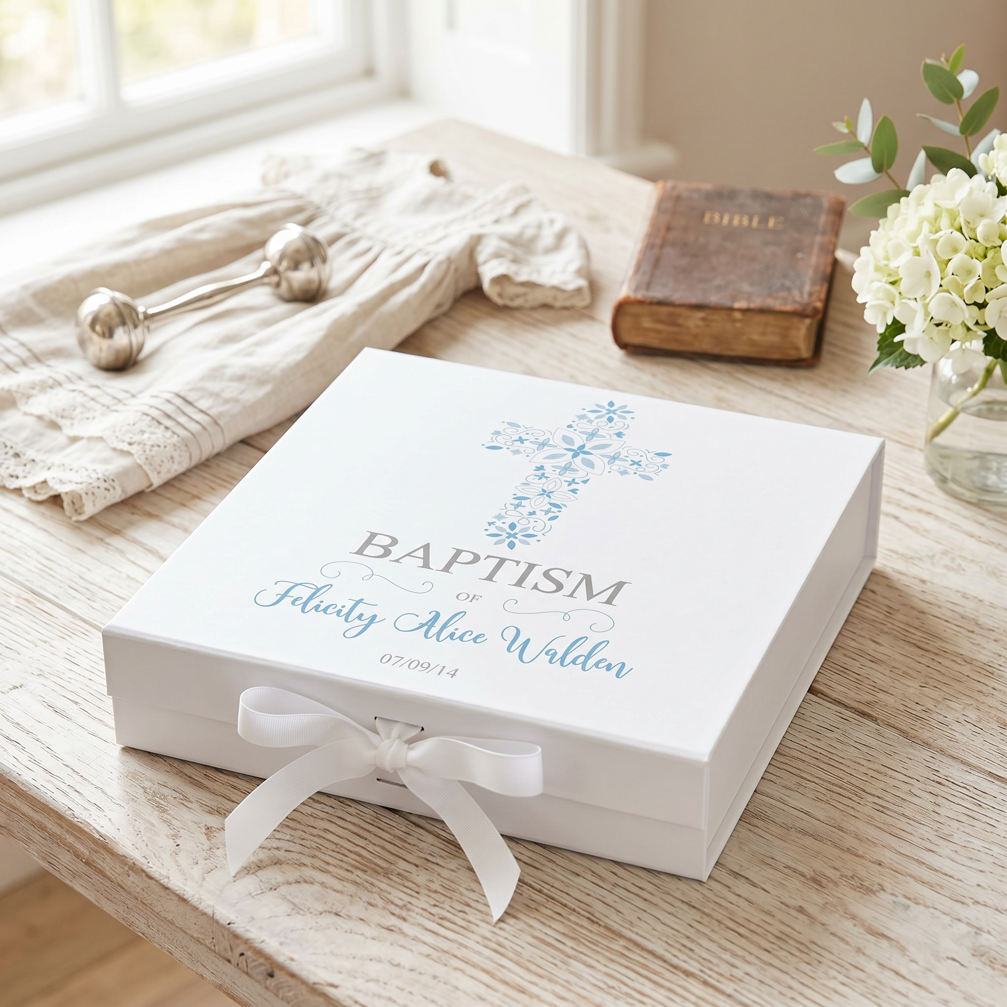 A white gift box with a blue cross and text, placed on a wooden table next to a vase of white flowers and a book.