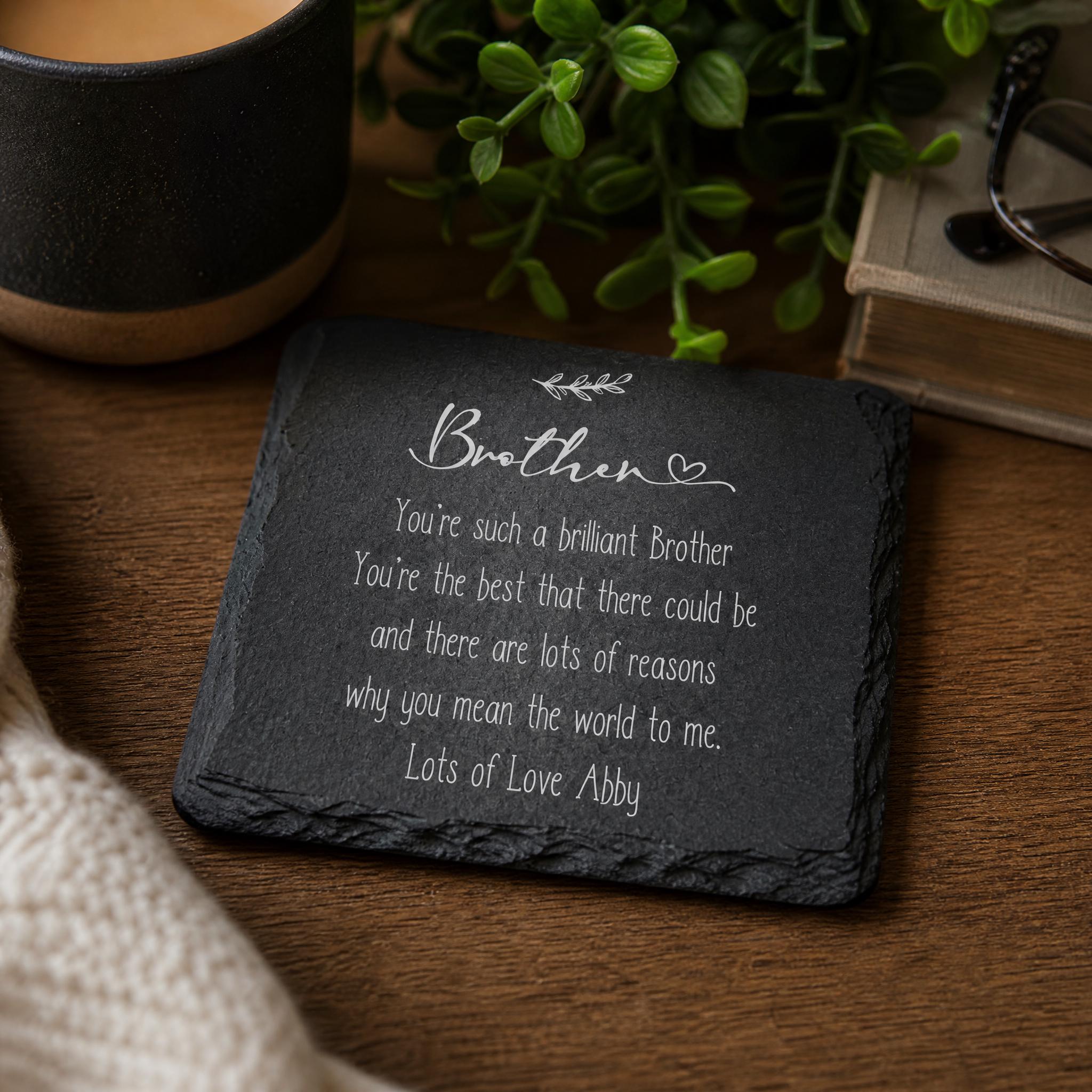A black slate coaster with a heartfelt message written on it, placed on a wooden surface next to a mug and a plant.