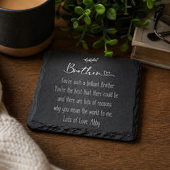 A black slate coaster with a heartfelt message written on it, placed on a wooden surface next to a mug and a plant.