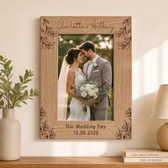 A wooden picture frame with a bride and groom photo inside, surrounded by decorative floral designs. The frame has the names "Charlotte & Anthony" and the date "Our Wedding Day 12.06.2026" engraved on it. The frame is placed on a shelf next to a lamp and a plant, creating a romantic and celebratory atmosphere.