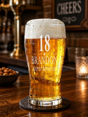 A personalized glass of beer with the number 18 and the name "Brandon" printed on it, placed on a wooden table in a bar setting.