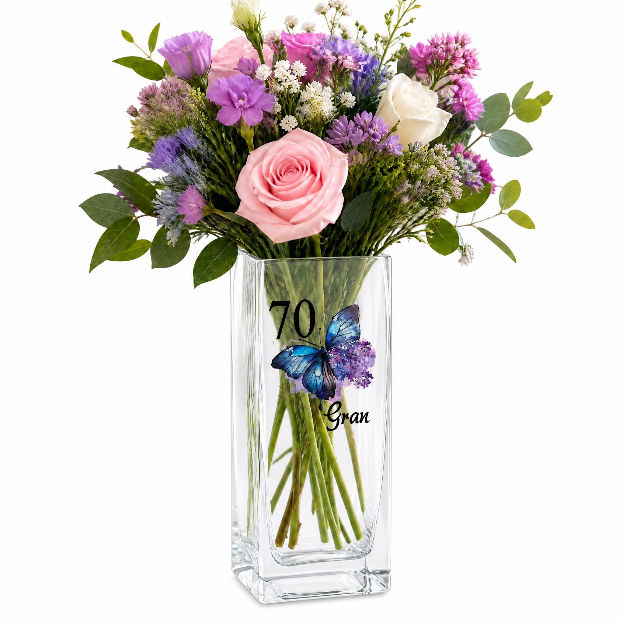 A clear glass vase filled with a bouquet of colorful flowers, including roses, lavender, and other blooms, along with green leaves.