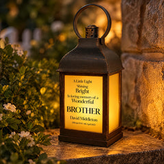 A black lantern with a message on it, placed on a stone surface next to some greenery.