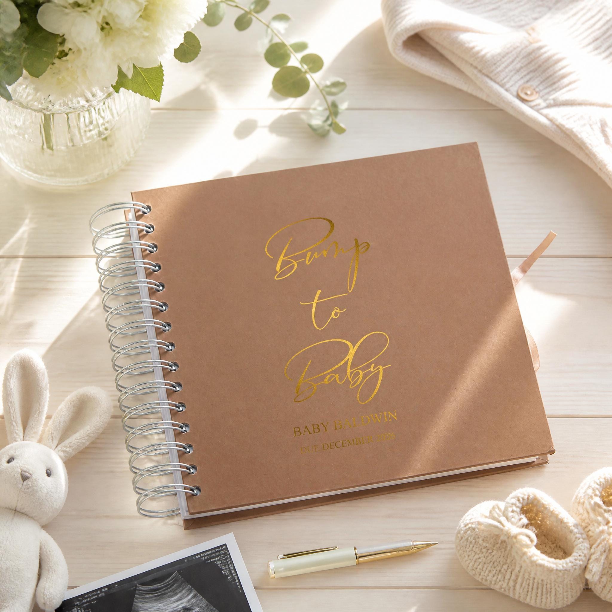 a brown notebook with the text "Bump to Baby" printed on it, along with the name "Baby Baldwin" and the date "DECEMBER 2016". The notebook is placed on a wooden surface, surrounded by various items such as a vase with flowers, a stuffed rabbit, and a pair of baby booties.