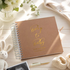 a brown notebook with the text "Bump to Baby" printed on it, along with the name "Baby Baldwin" and the date "DECEMBER 2016". The notebook is placed on a wooden surface, surrounded by various items such as a vase with flowers, a stuffed rabbit, and a pair of baby booties.