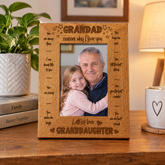 A wooden picture frame with a photo of a man and a young girl, likely a grandfather and granddaughter, inside it. The frame has a message engraved on it that says "GRANDDAD, reasons why I love you, let's always talk to each other, you are always funny to me, you make me laugh, you are my best friend". The frame is placed on a wooden table, with a potted plant and a book visible in the background.