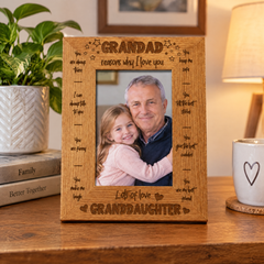 A wooden picture frame with a photo of a man and a young girl, likely a grandfather and granddaughter, inside it. The frame has a message engraved on it that says "GRANDDAD, reasons why I love you, let's always talk to each other, you are always funny to me, you make me laugh, you are my best friend". The frame is placed on a wooden table, with a potted plant and a book visible in the background.